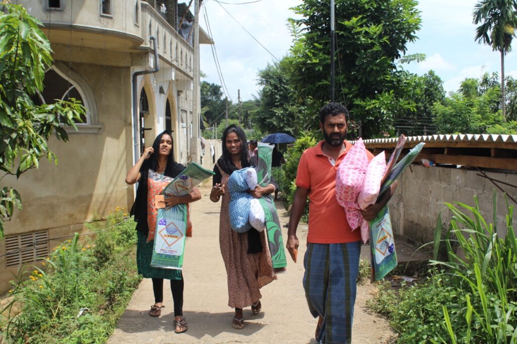 Three people carrying sleeping mats and pillows along a village path