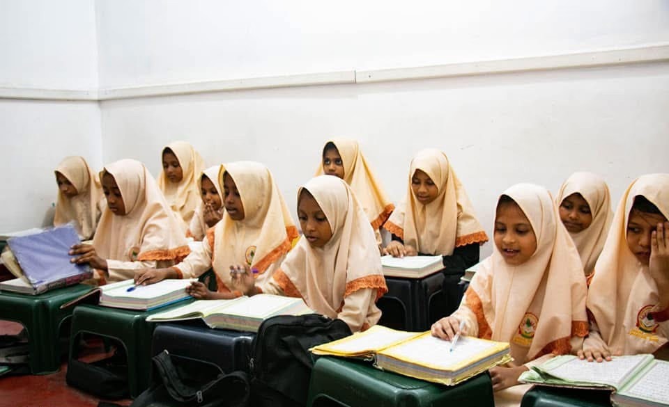 Schoolgirls in pale headscarves writing in exercise books at their desks in a classroom