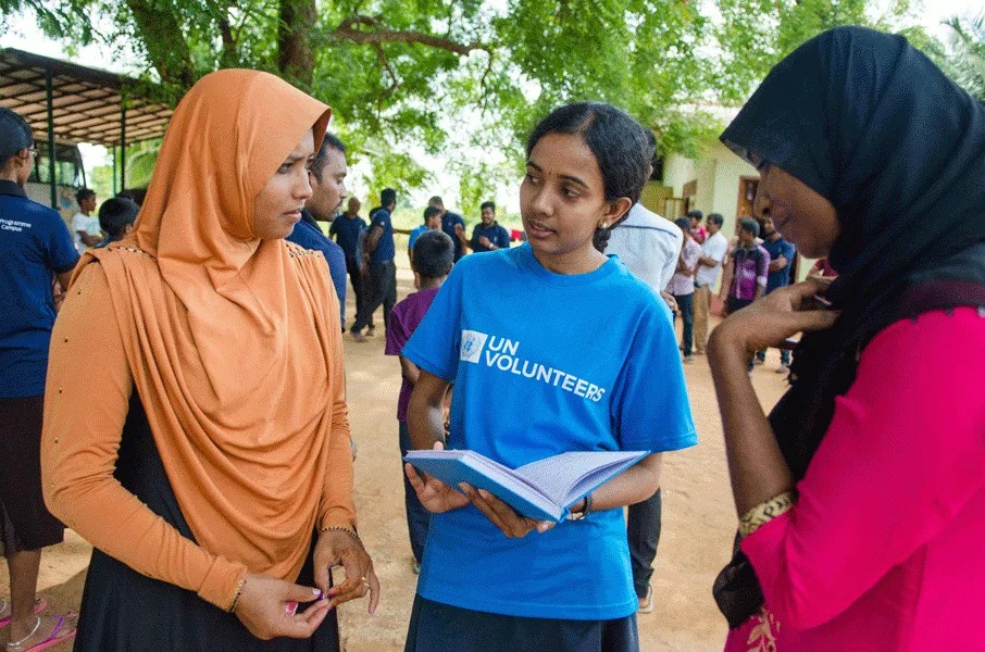 A volunteer wearing a UN Volunteers t-shirt speaking with two women outdoors during a community visit