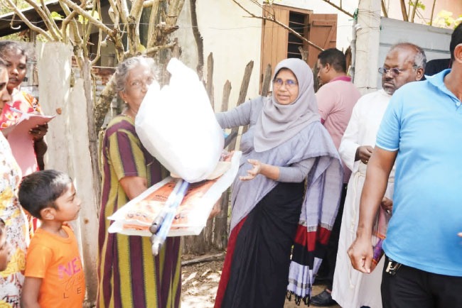 A woman passing a large sack of dry food rations to an older woman outside a house, with neighbours gathered around