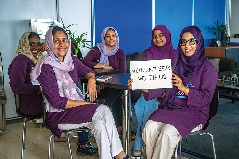 A group of volunteers seated around a table, one holding a handwritten sign reading Volunteer With Us